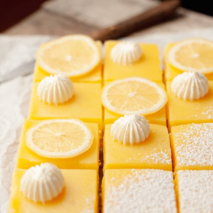 gluten free lemon bars with a shortbread crust topped with powdered sugar, whipped cream and slices of lemons on parchment paper