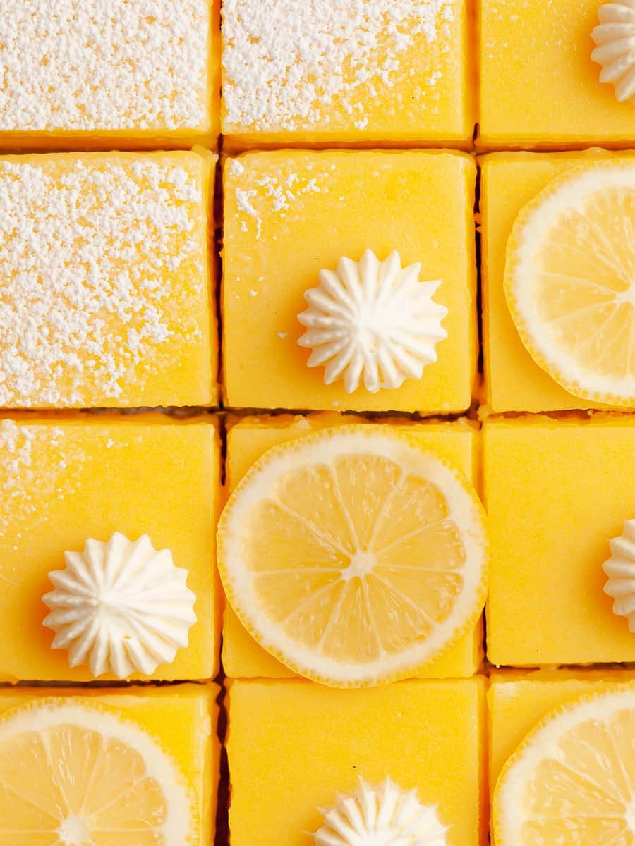 gluten free lemon bars with a shortbread crust topped with powdered sugar, whipped cream and slices of lemons on parchment paper