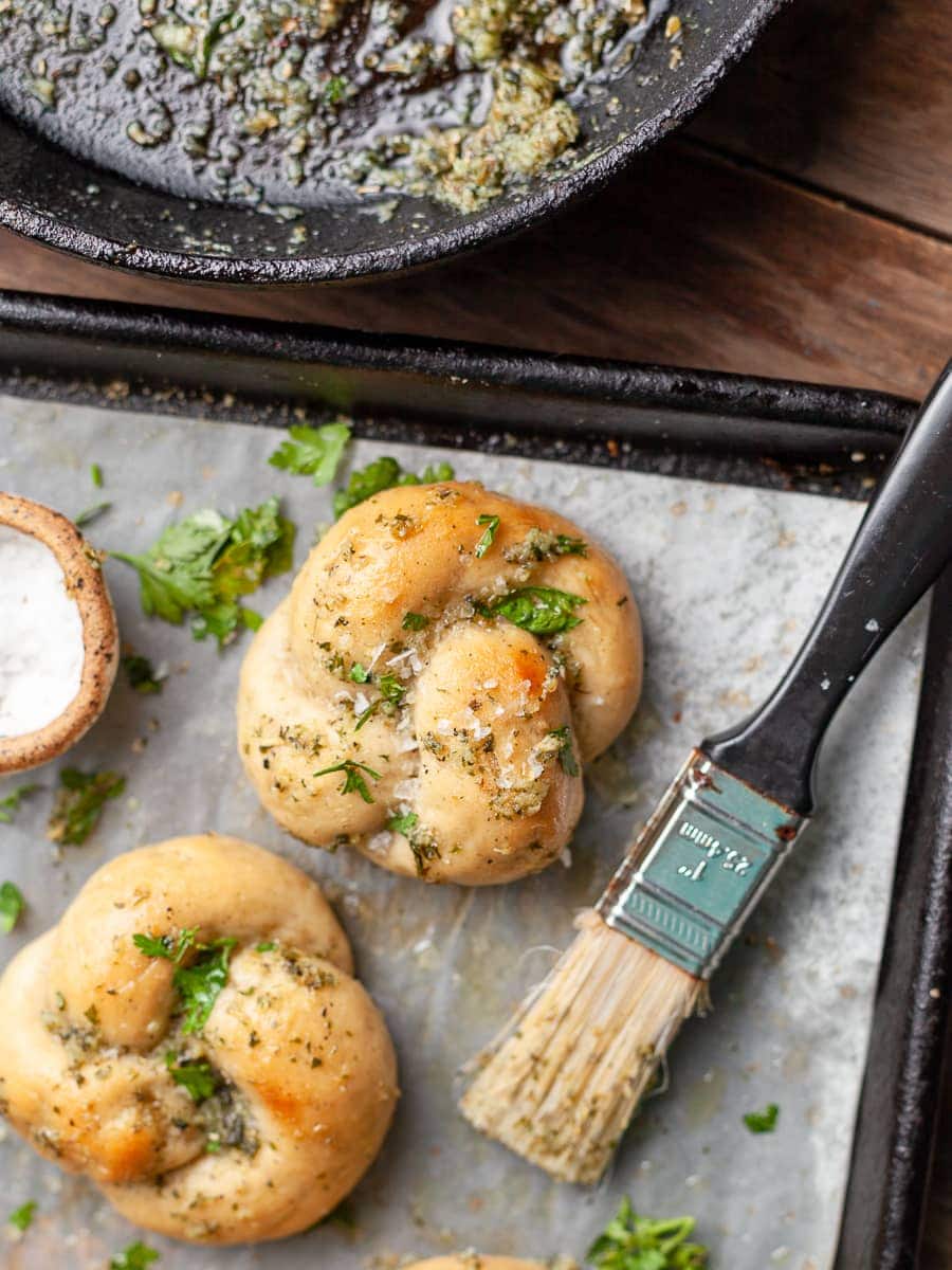 gluten free garlic knots on a sheet tray, topped with garlic butter, next to it is a used pastry brush and a bowl of flaky salt