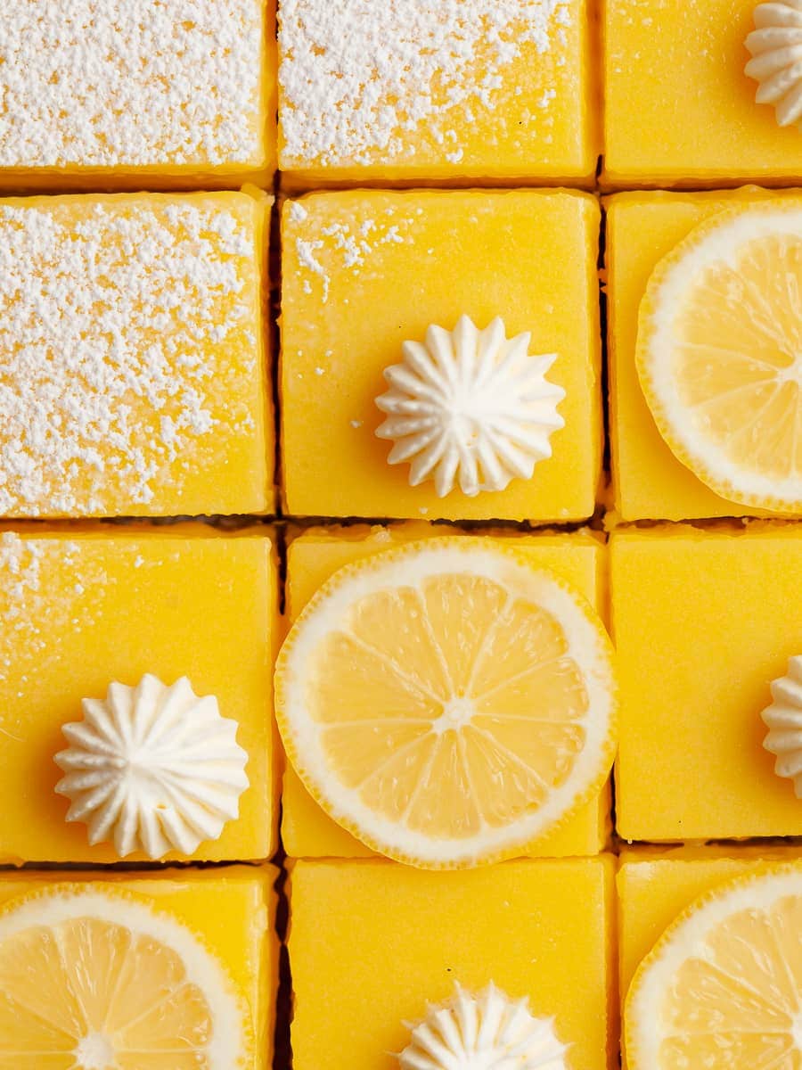 gluten free lemon squares topped with powdered sugar, whipped cream and slices of lemons on parchment paper