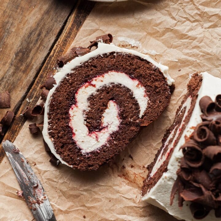 a slice of gluten free chocolate swiss roll filled with cherry jam and whipped cream. topped with whipped cream and chocolate curls