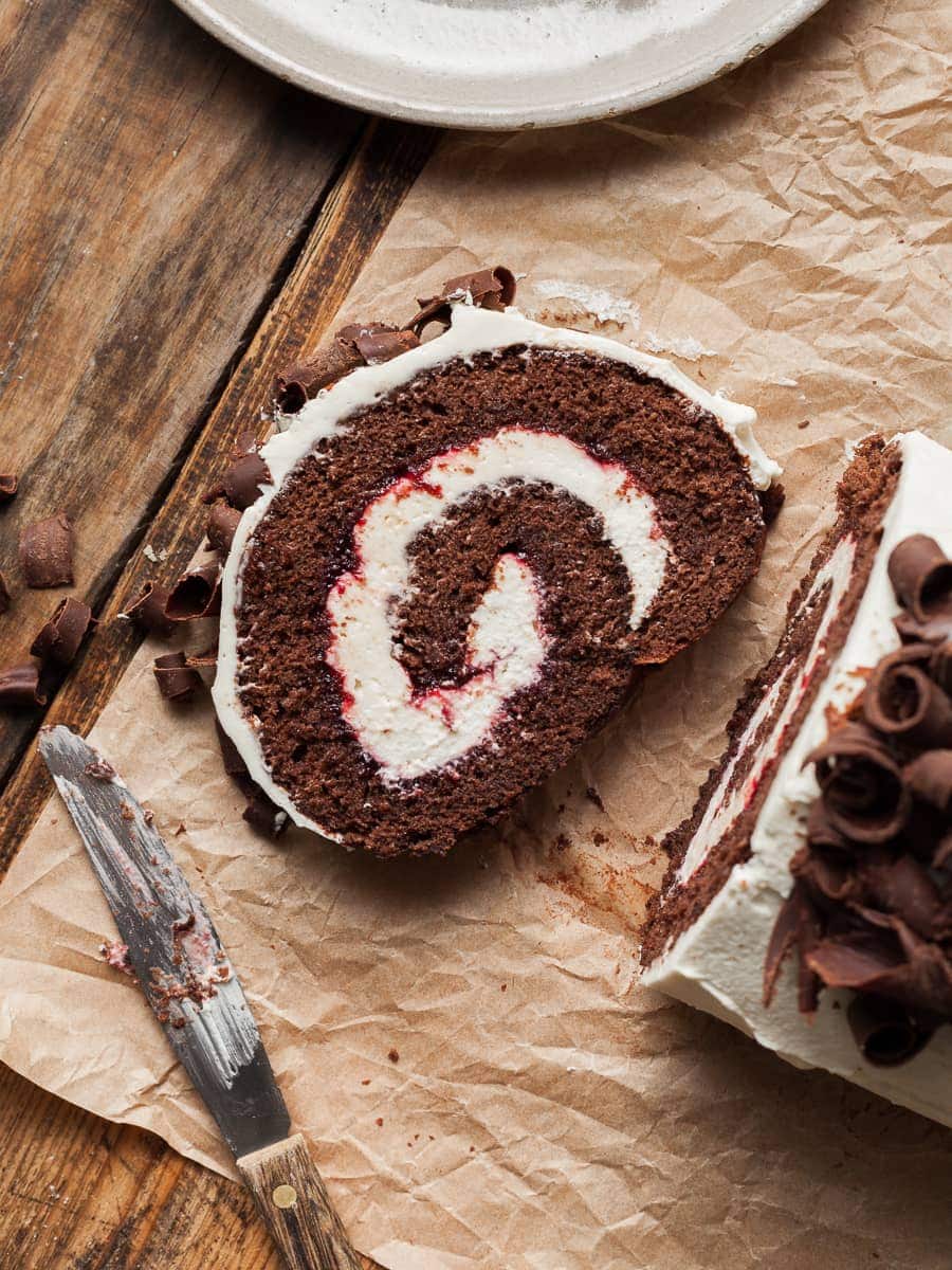 a slice of gluten free chocolate swiss roll filled with cherry jam and whipped cream. topped with whipped cream and chocolate curls