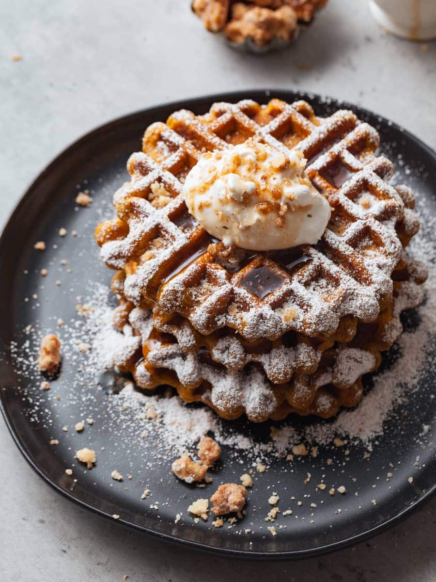 a stack of pumpkin waffles topped with powdered sugar, whipped cream and crushed walnuts