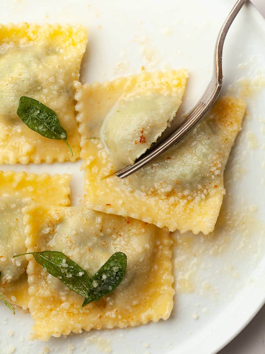 gluten free spinach ricotta ravioli on a white plate with brown butter, parmesan cheese and crispy sage, a fork in the corner that's splitting one ravioli in half